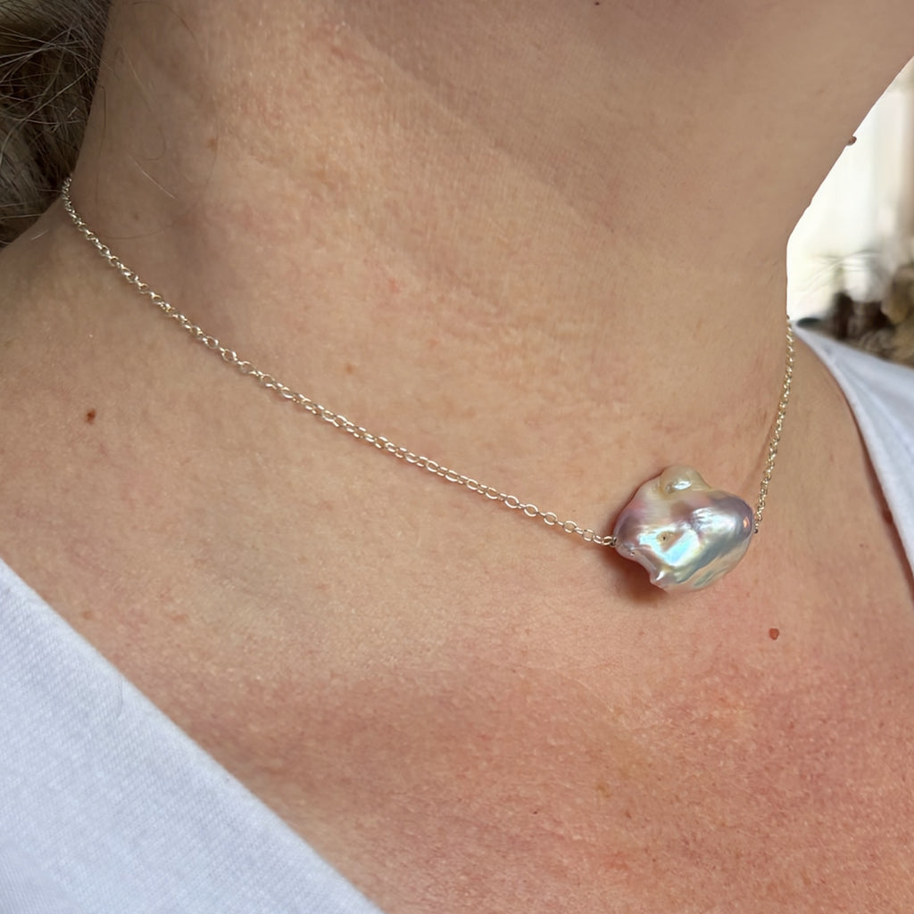 Angled close-up of a pink Keshi pearl necklace worn at the collarbone, showing the pearl’s irregular contours and bright cream, mauve and blue-green lustre on a fine chain.