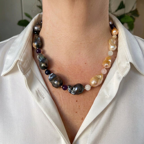 Statement necklace with dark blue-grey keshi pearls along one side and golden-cream keshi pearls along the other, separated by small purple, navy and pale pink gemstone beads, worn with an open-neck white shirt.