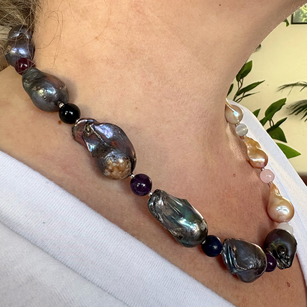 Side-angle view of large iridescent dark blue-grey and warm cream keshi pearls separated by small multicoloured gemstone beads, worn as a statement necklace with a white V-neck top.