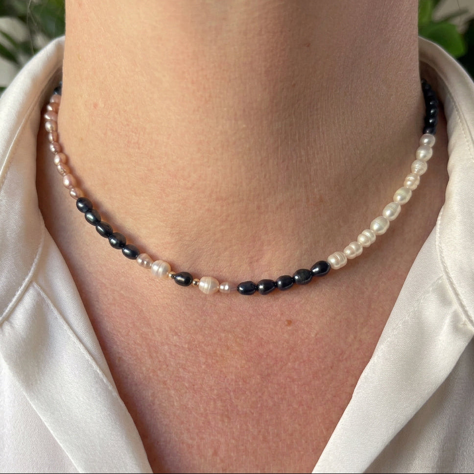 Blush-pink, cream and dark blue rice pearls arranged in sections along a delicate necklace with small silver-coloured spacers, worn at the collarbone with an open-neck white shirt.