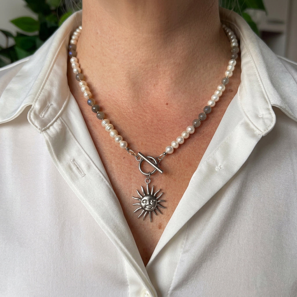 White baroque pearl and grey labradorite necklace with a fastened front toggle and detailed stainless steel sun face pendant, worn with an open-neck white shirt.