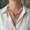 White baroque pearl necklace with blue-green glass beads, a fastened front toggle and a detailed stainless steel dragonfly pendant, worn with an open-neck white shirt.