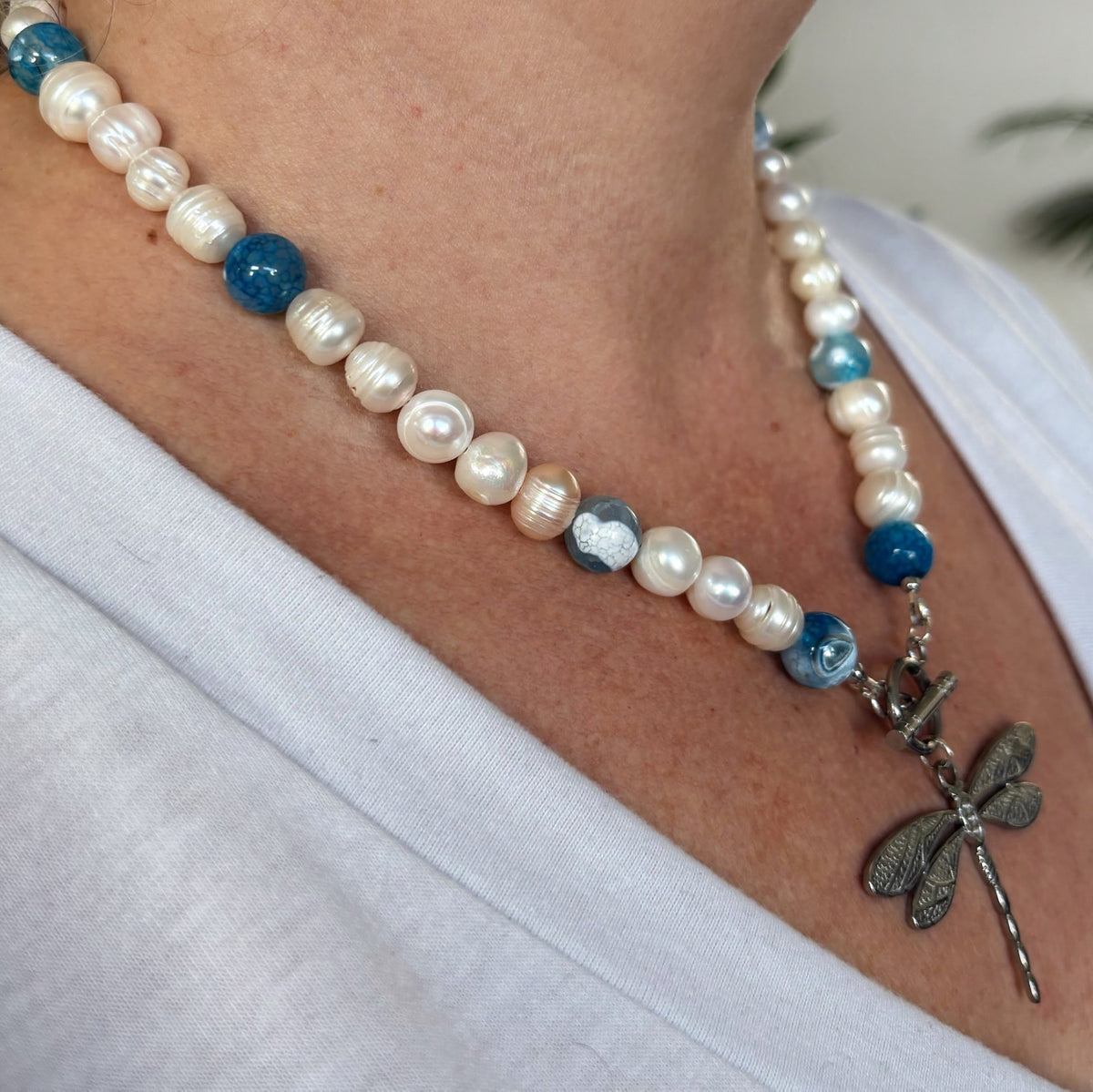 Angled close-up of a white baroque pearl necklace worn with a white top, showing naturally ridged pearls, blue glass accent beads and a stainless steel dragonfly pendant below the front toggle clasp.