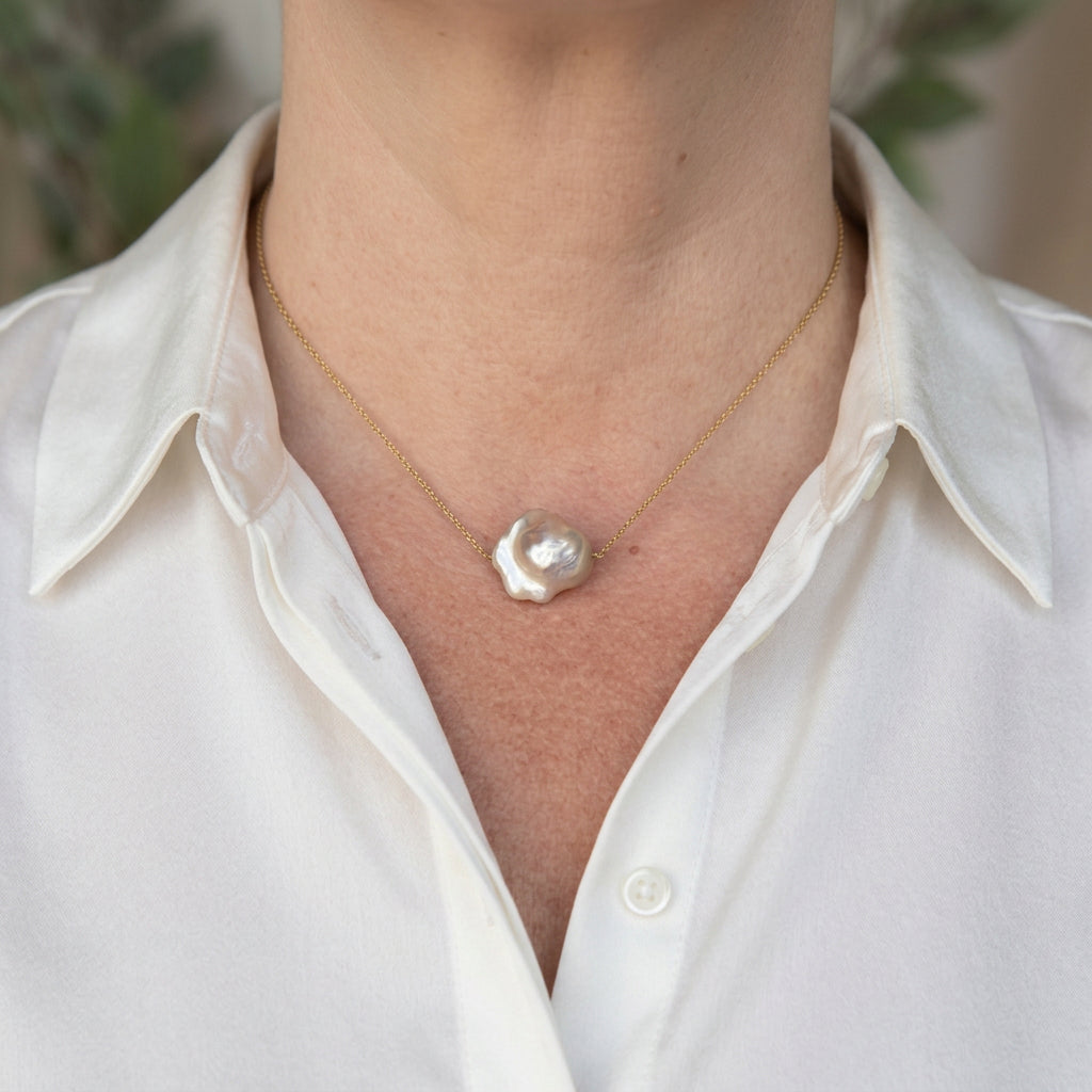 Pink keshi pearl necklace on a delicate gold-filled chain, worn with an open-neck white shirt, showing an irregular petal-shaped pearl with blush tones and bright silvery lustre.