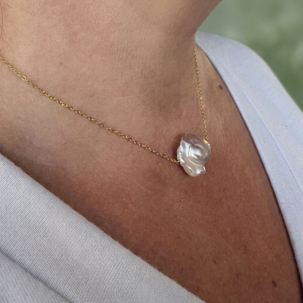 Angled close-up of a pink Keshi pearl necklace worn with a white V-neck top, showing the pearl’s folded, petal-like shape and creamy pink lustre on a fine gold-filled chain.