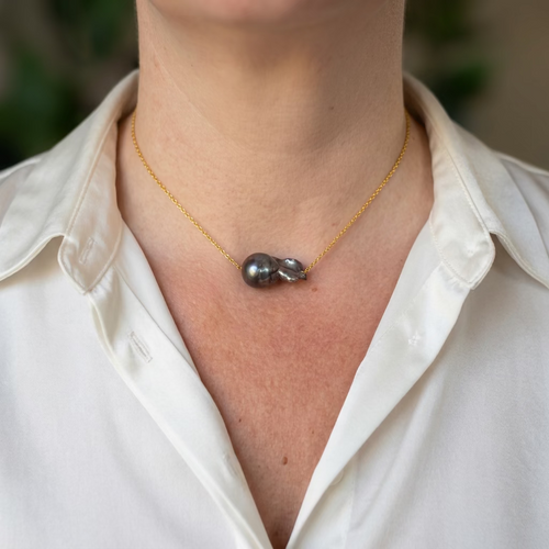 Large dark blue keshi pearl on a delicate gold-filled chain, worn with an open-neck white shirt, showing a rounded dark dome and folded nacre with silvery-purple highlights.