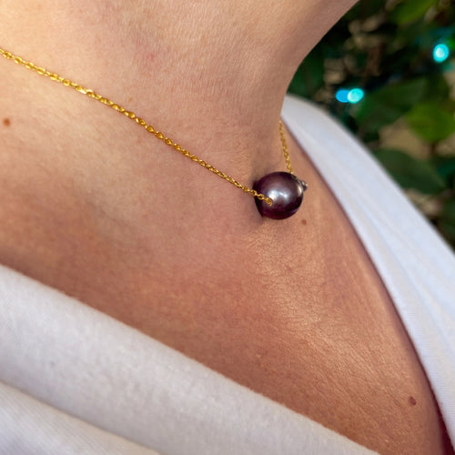 Side-angle view of a dark blue keshi pearl on a fine gold-filled chain, worn with a white V-neck top, highlighting the pearl’s glossy rounded dome and deep violet iridescence.