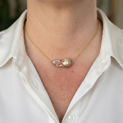 Large rainbow keshi pearl on a delicate gold-filled chain, worn with an open-neck white shirt, showing a rounded champagne-gold end and rippled pink-lavender nacre.