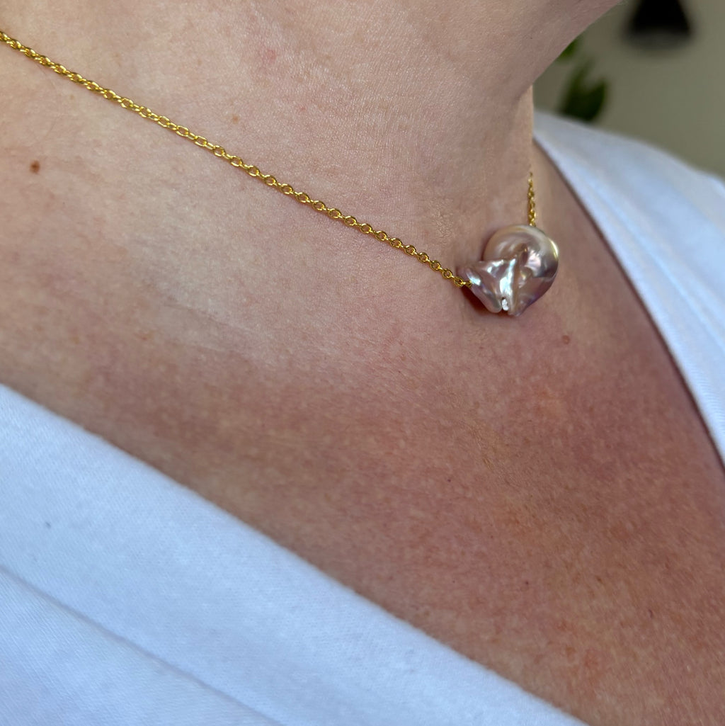 Side-angle view of a rainbow keshi pearl on a fine gold-filled chain, worn with a white V-neck top, showing the pearl’s sculptural folds and rose-silver iridescence.⠋