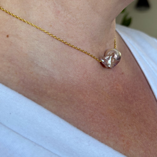 Side-angle view of a rainbow keshi pearl on a fine gold-filled chain, worn with a white V-neck top, showing the pearl’s sculptural folds and rose-silver iridescence.⠋