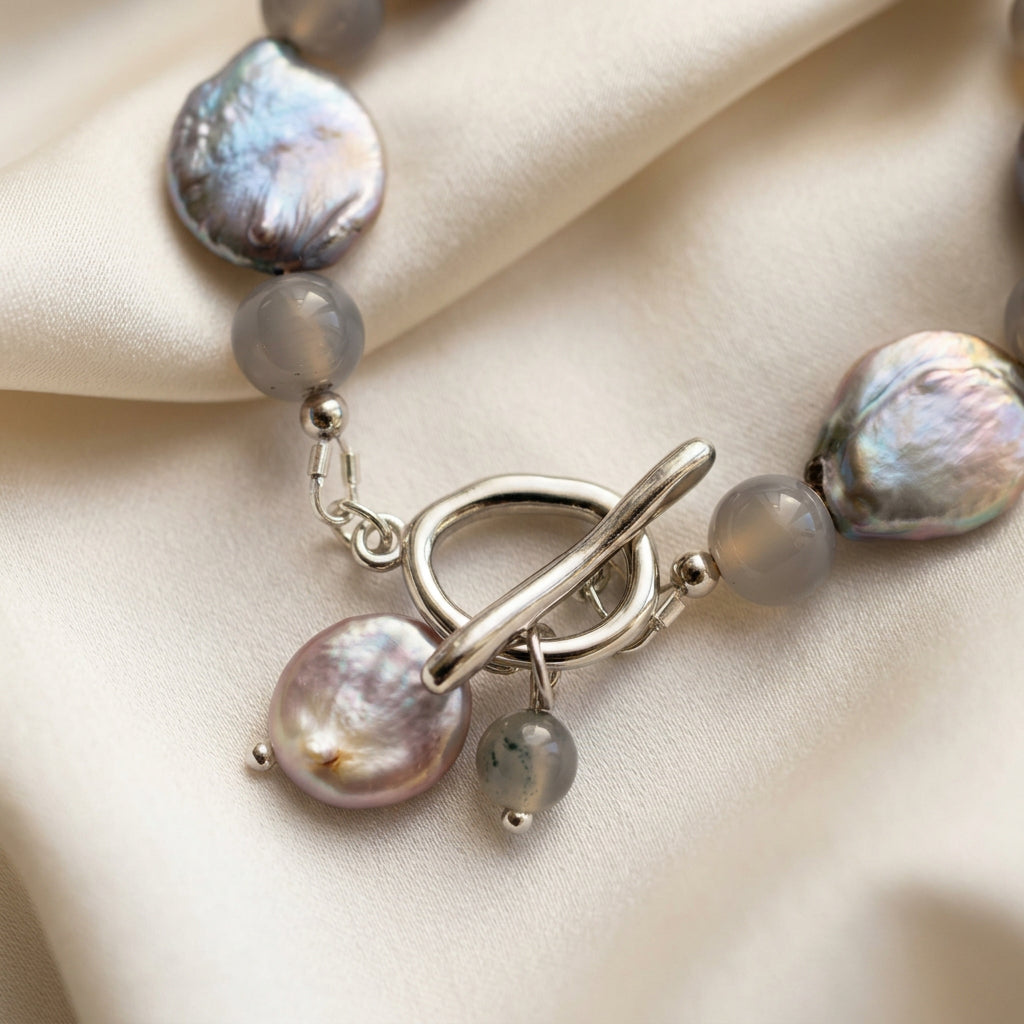 Close-up of a fastened stainless steel toggle clasp with dangling coin pearl and grey labradorite charms, framed by iridescent silver-toned coin pearls and round labradorite beads on cream satin.