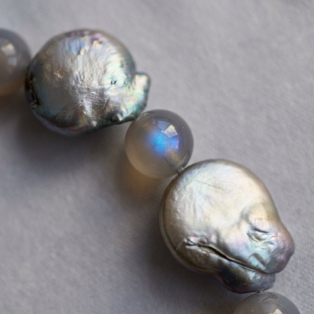 Macro close-up of a grey labradorite bead with a vivid blue flash between iridescent silver-toned coin pearls, showing their irregular outlines and naturally rippled surfaces on pale fabric.⠋