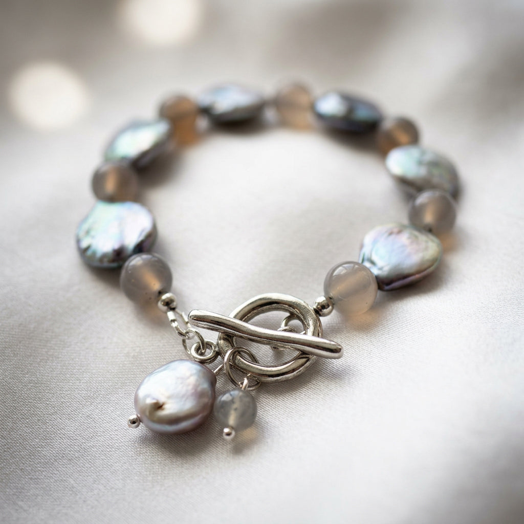 Labradorite and silver-toned coin pearl bracelet arranged in an oval on cream satin, with the fastened stainless steel toggle clasp and dangling pearl and labradorite charms in the foreground.