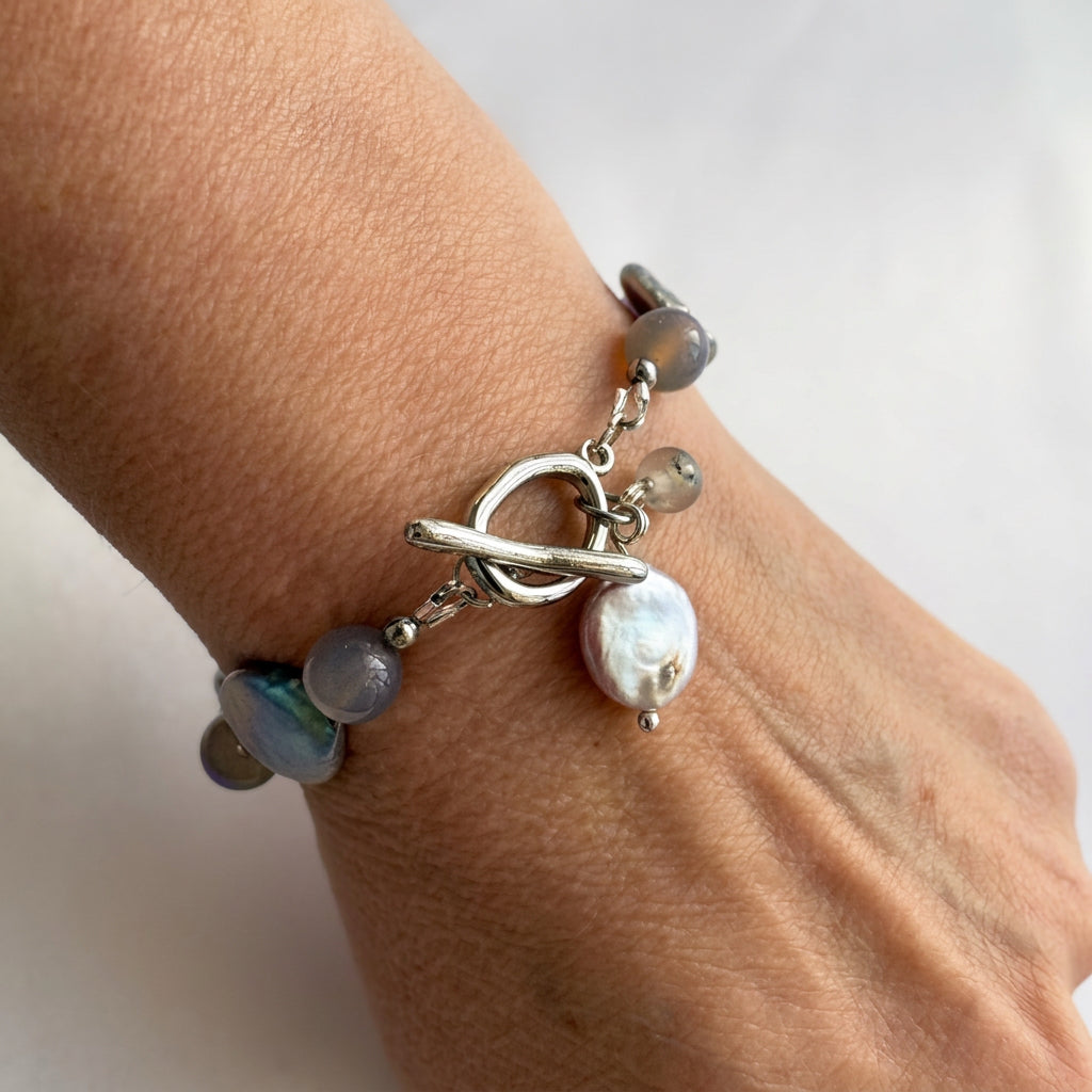Close-up of a labradorite and silver-toned coin pearl bracelet worn on a wrist, highlighting the fastened stainless steel toggle clasp and dangling iridescent pearl and grey labradorite charms.