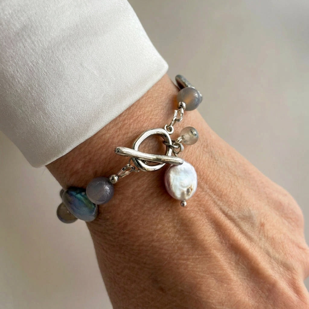Grey labradorite and iridescent coin pearl bracelet with a fastened stainless steel toggle clasp and dangling pearl and gemstone charms, worn beside a white silk shirt cuff.