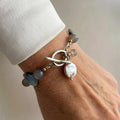 Grey labradorite and iridescent coin pearl bracelet with a fastened stainless steel toggle clasp and dangling pearl and gemstone charms, worn beside a white silk shirt cuff.