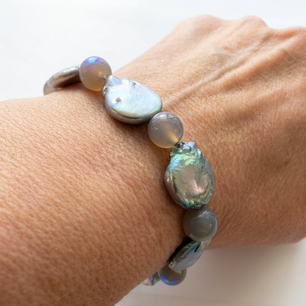 Close-up of a bracelet worn on a wrist, with iridescent silver-toned coin pearls alternating with round grey labradorite beads showing flashes of blue and gold.⠋