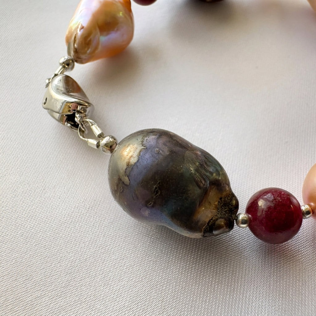 Close-up of a large dark keshi pearl with purple, green and bronze iridescence beside a deep red garnet bead and small sterling silver accents, with the box clasp behind it on cream satin.
