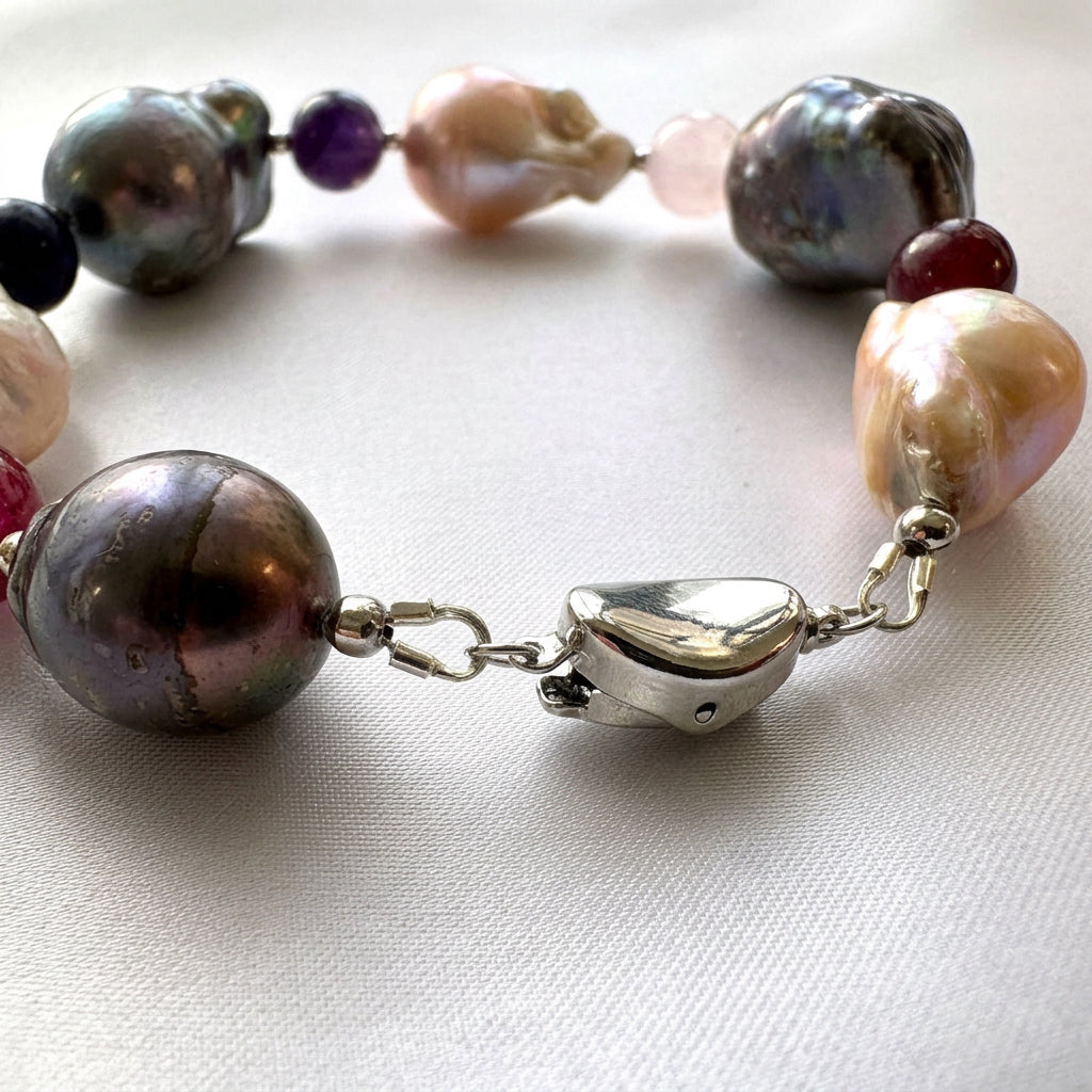 Close-up of a fastened polished sterling silver box clasp between large dark iridescent and blush-cream keshi pearls, with colourful gemstone beads behind it on cream satin.