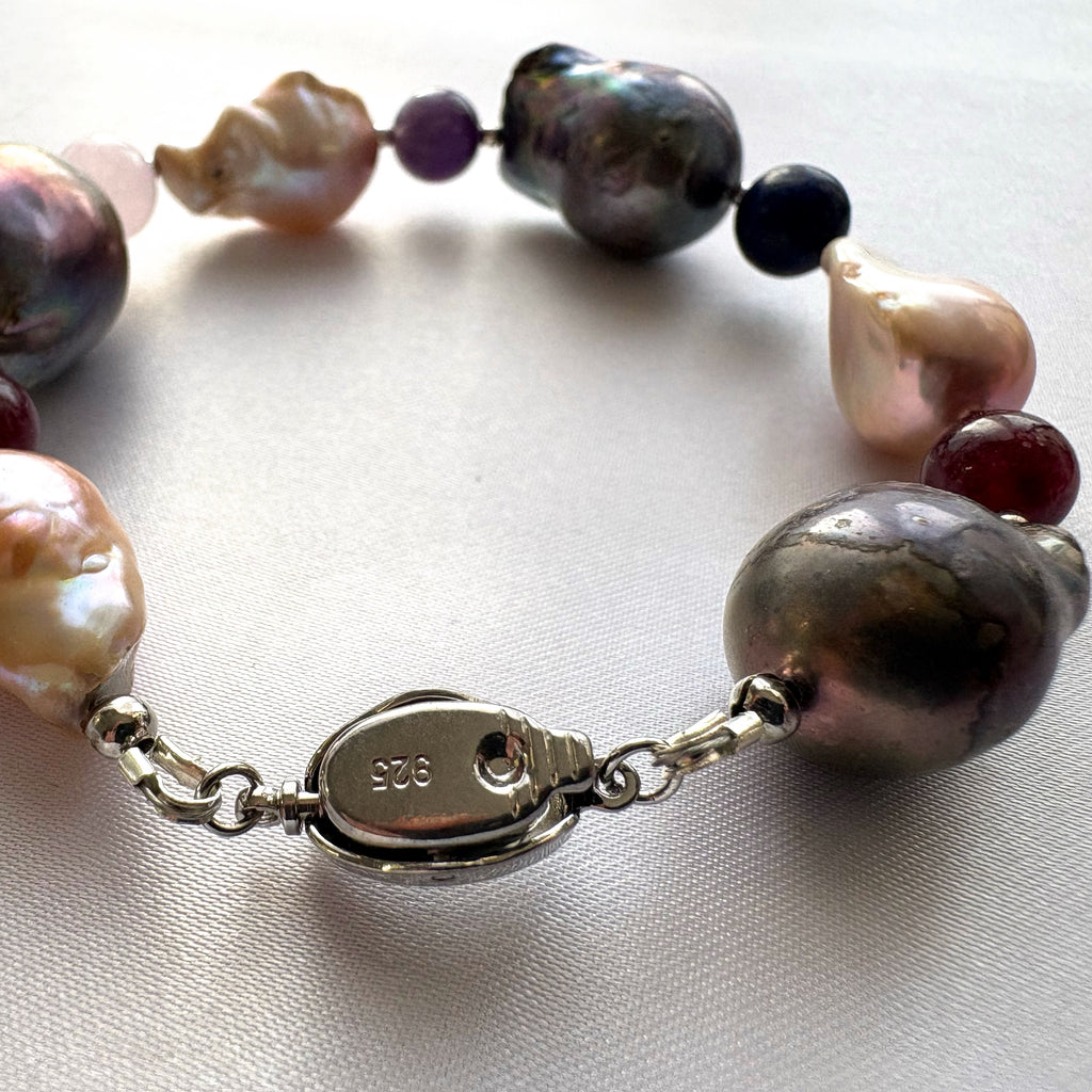 Close-up of the underside of a fastened sterling silver box clasp stamped 925, with large dark iridescent and blush-cream keshi pearls and colourful gemstone beads arranged on cream satin.