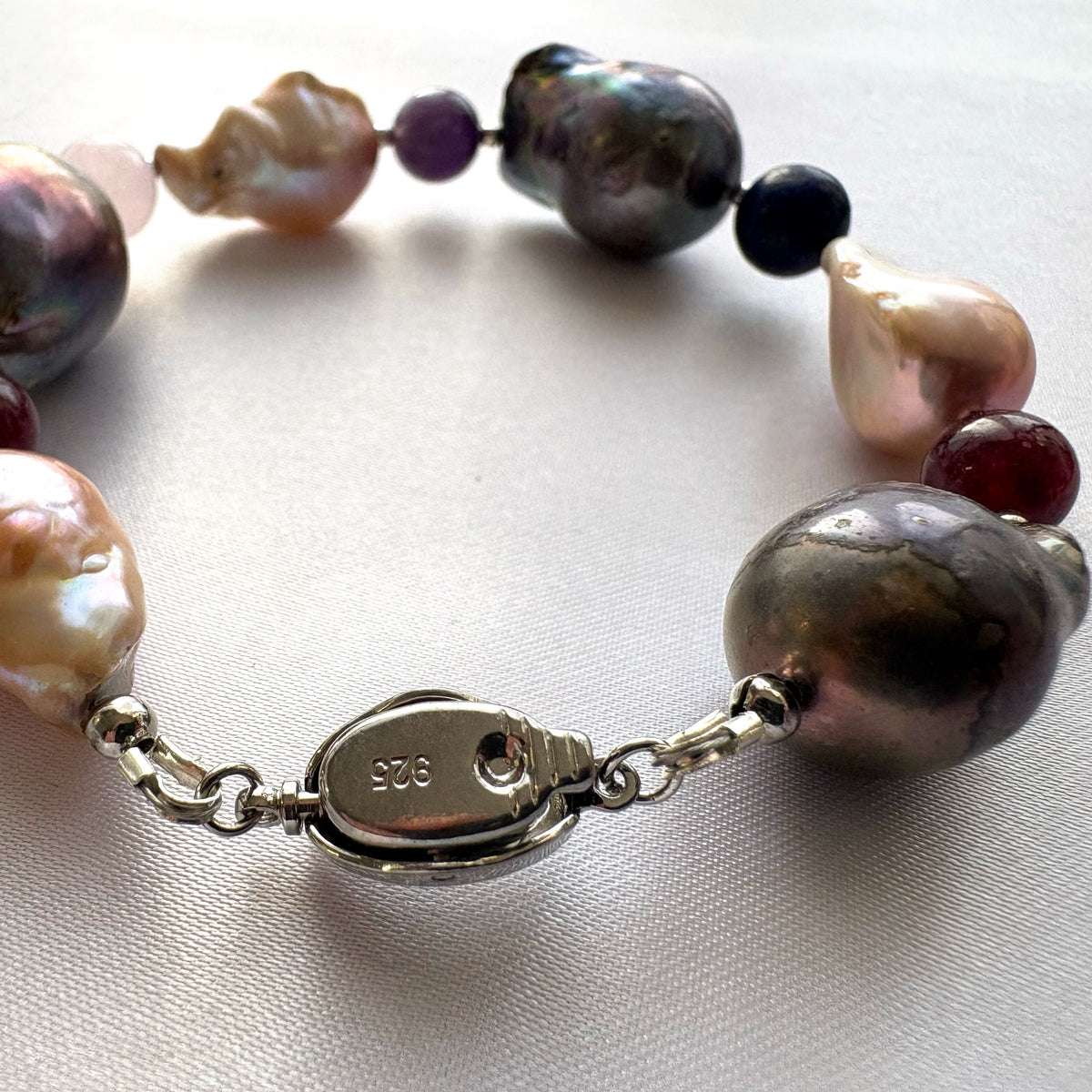 Close-up of the underside of a fastened sterling silver box clasp stamped 925, with large dark iridescent and blush-cream keshi pearls and colourful gemstone beads arranged on cream satin.