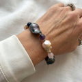 Large dark iridescent and blush-cream keshi pearls separated by purple, pale pink and deep blue gemstone beads, worn as a bracelet beside a white silk shirt cuff.