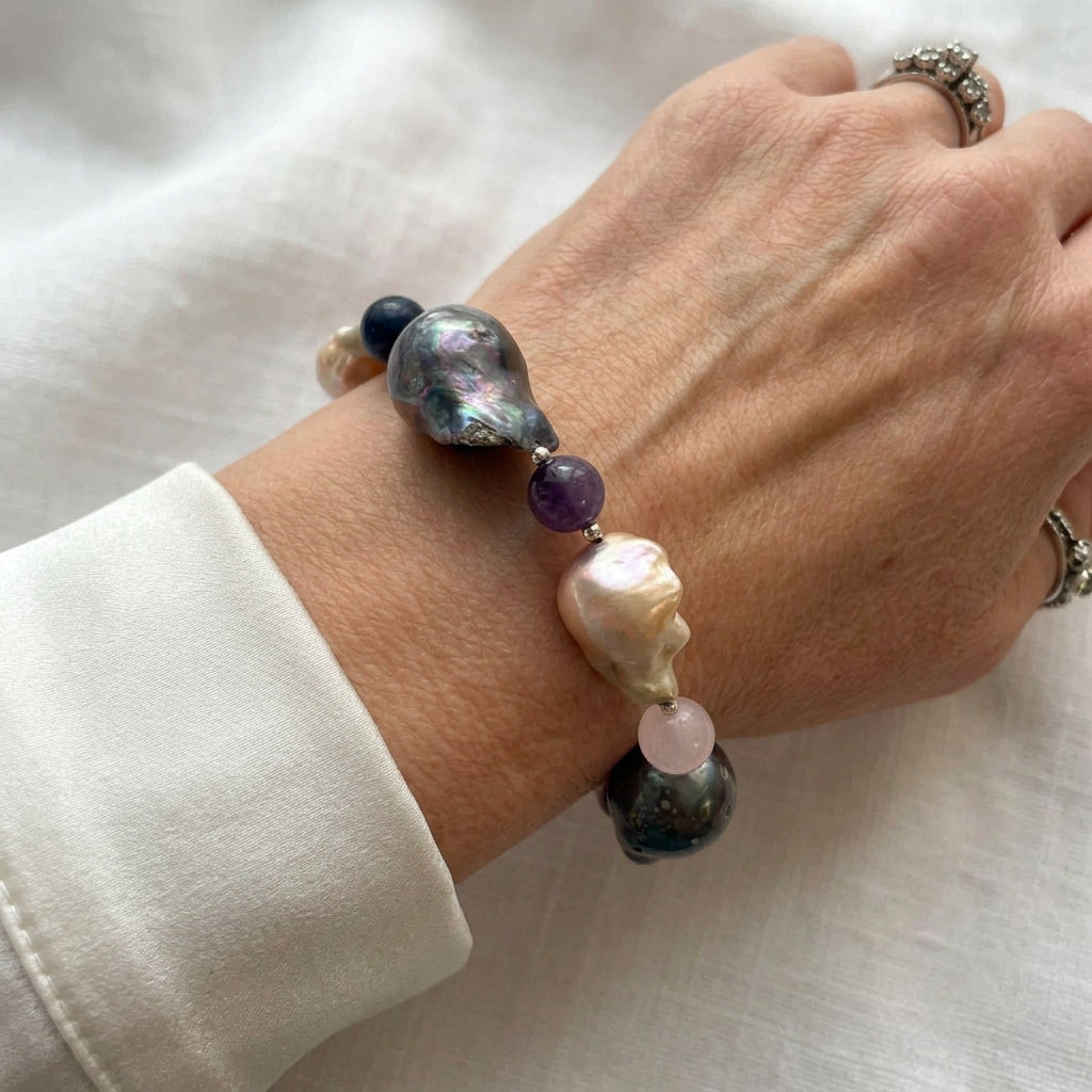 Large dark iridescent and blush-cream keshi pearls separated by purple, pale pink and deep blue gemstone beads, worn as a bracelet beside a white silk shirt cuff.