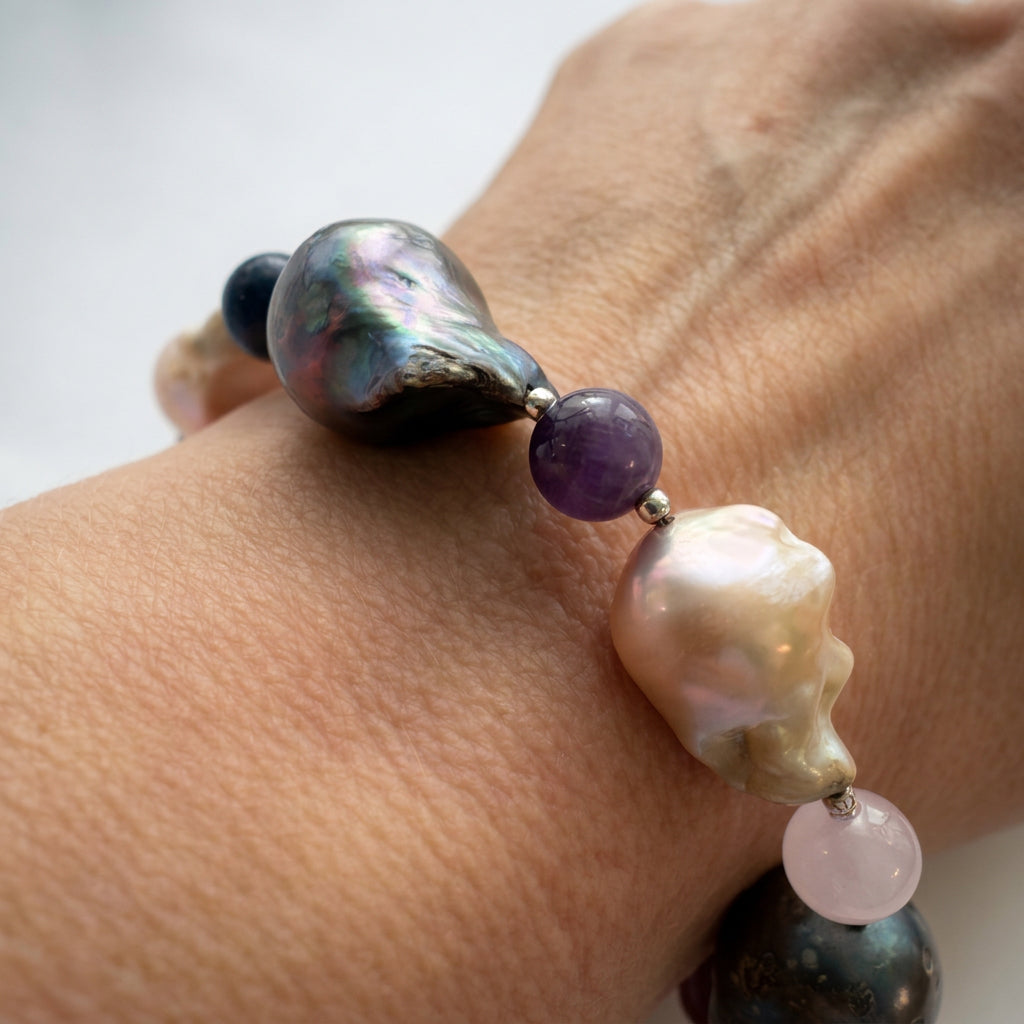 Close-up of a keshi pearl bracelet worn on a wrist, showing a large dark pearl with green-purple iridescence, a creamy blush pearl, purple amethyst, pale pink quartz and small sterling silver spacers.