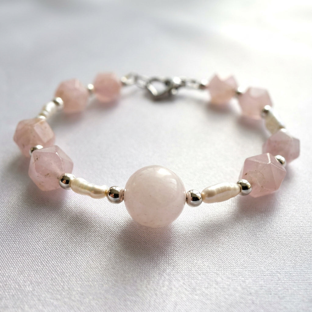 Pink quartz and freshwater pearl bracelet arranged in a circle on pale fabric, with a large round pink centre bead, faceted quartz beads, white rice-shaped pearls, silver-coloured hematite spacers and a heart clasp.