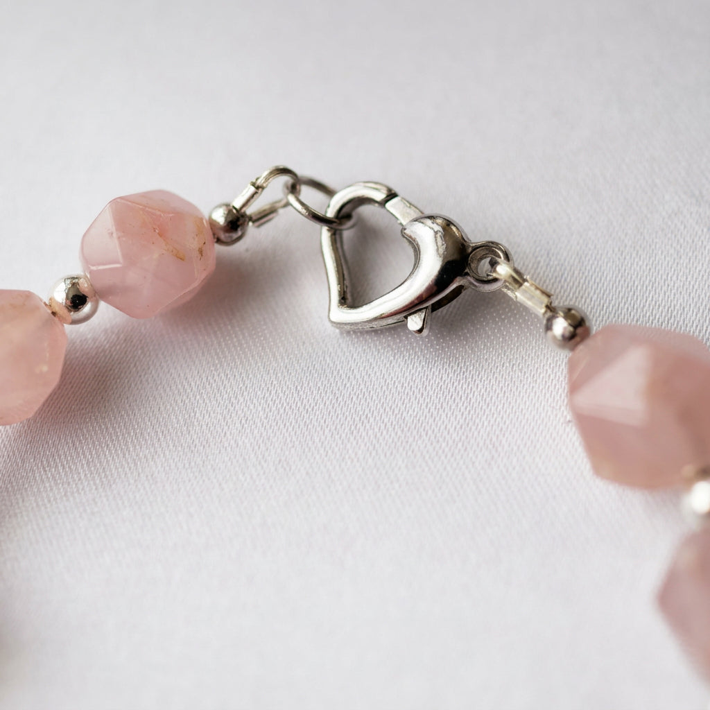 Close-up of the fastened silver-finished brass heart clasp between faceted pink quartz beads and small silver-coloured hematite spacers on white fabric.