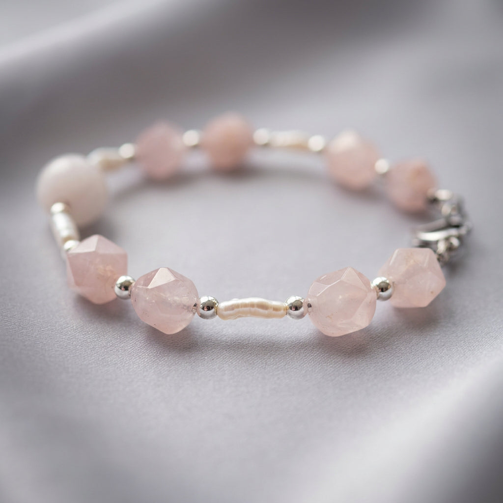 Pink quartz and freshwater pearl bracelet resting in a circle on grey fabric, with a white rice-shaped pearl between faceted blush-pink beads and polished silver-coloured hematite spacers in the foreground.⠋