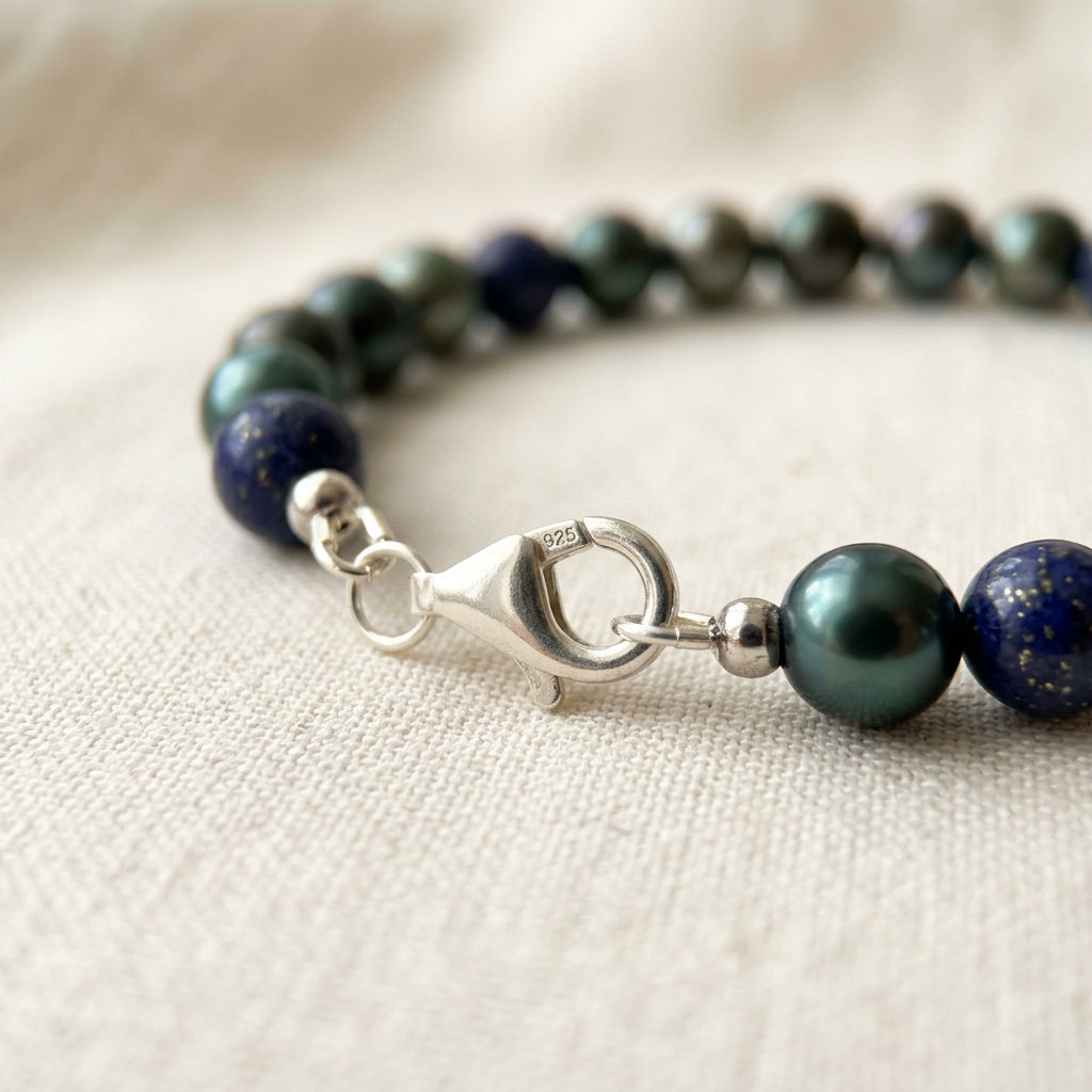 Close-up of a fastened sterling silver lobster clasp stamped 925, connecting deep green peacock pearls and navy-blue lapis lazuli beads on cream linen.