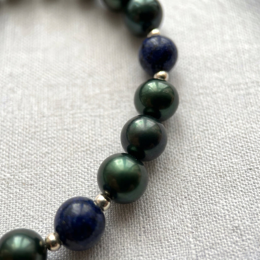 Close-up of three lustrous deep green peacock pearls between navy-blue lapis lazuli beads and small sterling silver spacers on pale linen.