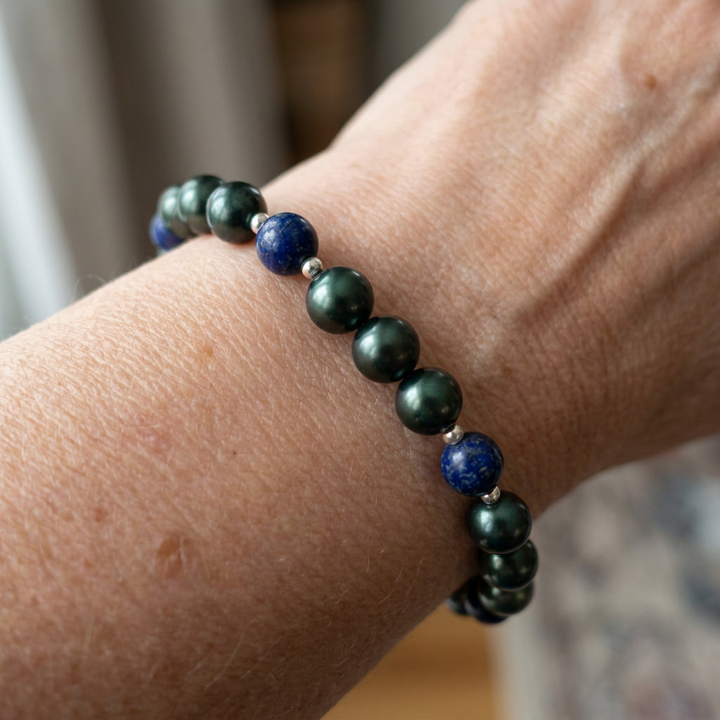 Angled view of a peacock pearl and lapis lazuli bracelet worn on a wrist, showing groups of deep green pearls between navy-blue beads with small sterling silver spacers against a white background.