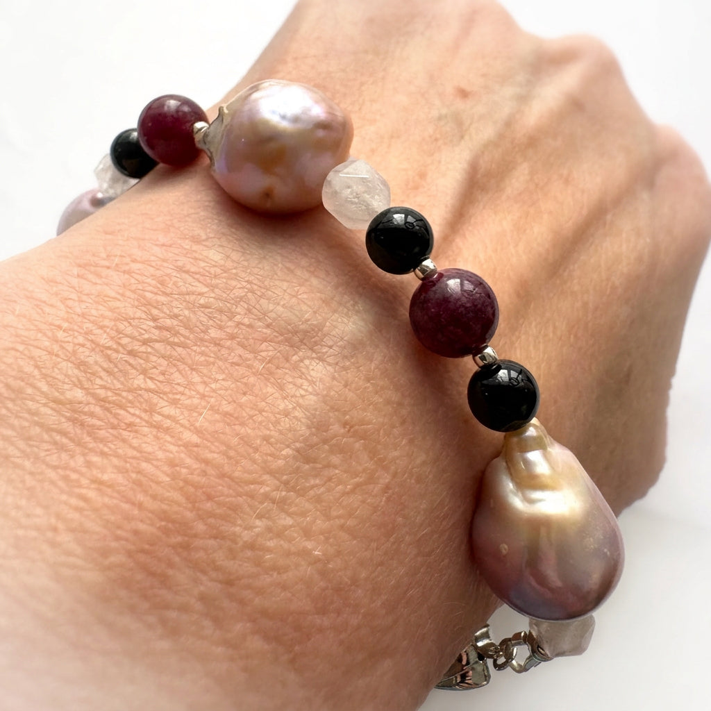 Close-up of a keshi pearl bracelet worn on a wrist, showing large blush-cream pearls with lavender lustre, deep red garnet between glossy black onyx beads, faceted pale quartz and small sterling silver accents.