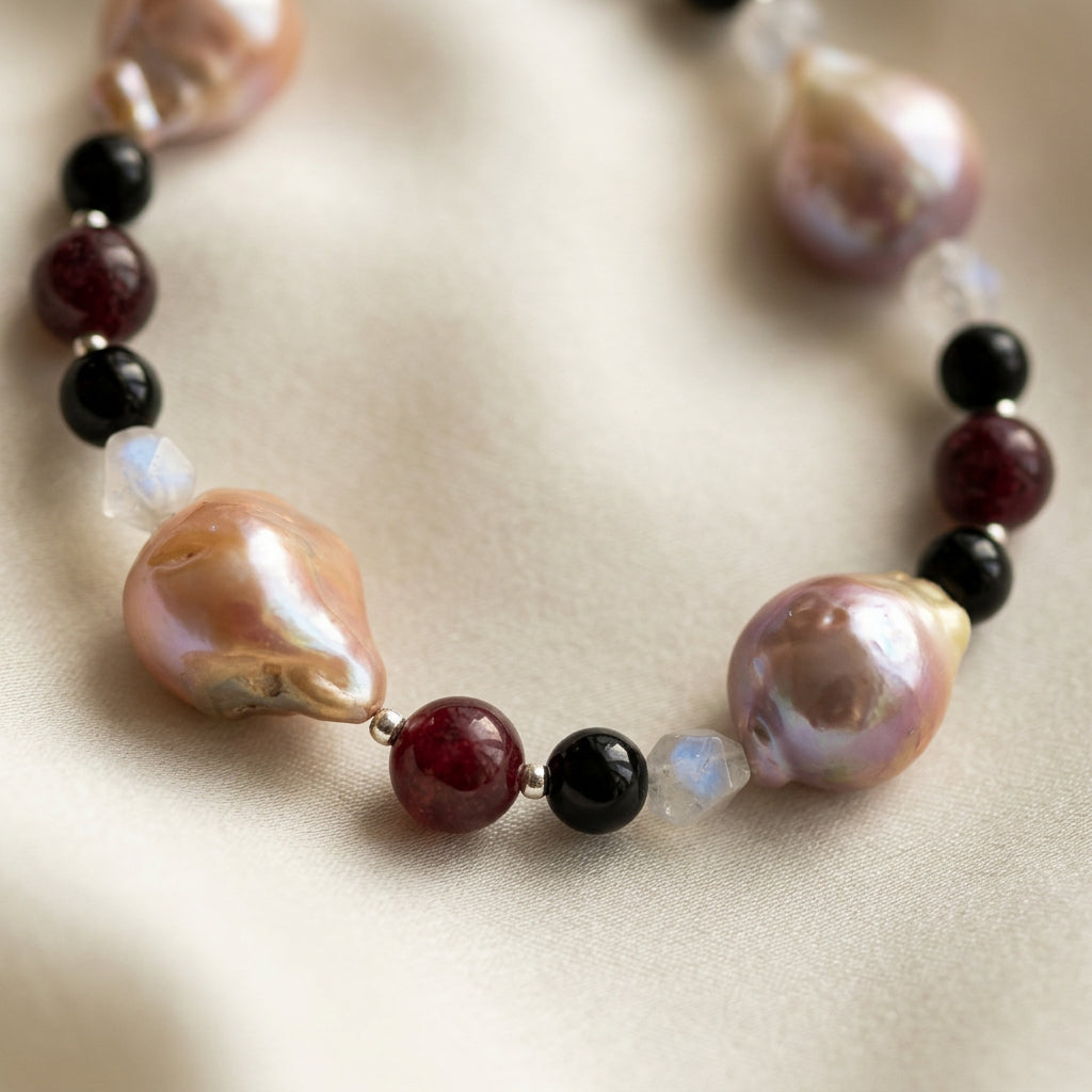 Close-up of blush-pink and peach keshi pearls with lavender iridescence, interspersed with deep red garnet, glossy black onyx, faceted pale quartz and small sterling silver beads on cream satin.