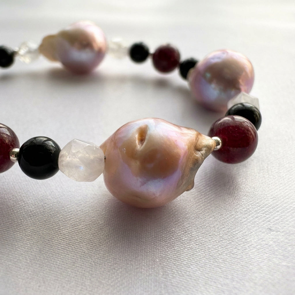 Close-up of a large irregular blush-cream keshi pearl with pink and lavender iridescence, beside faceted pale quartz, glossy black onyx and deep red garnet beads on cream satin.