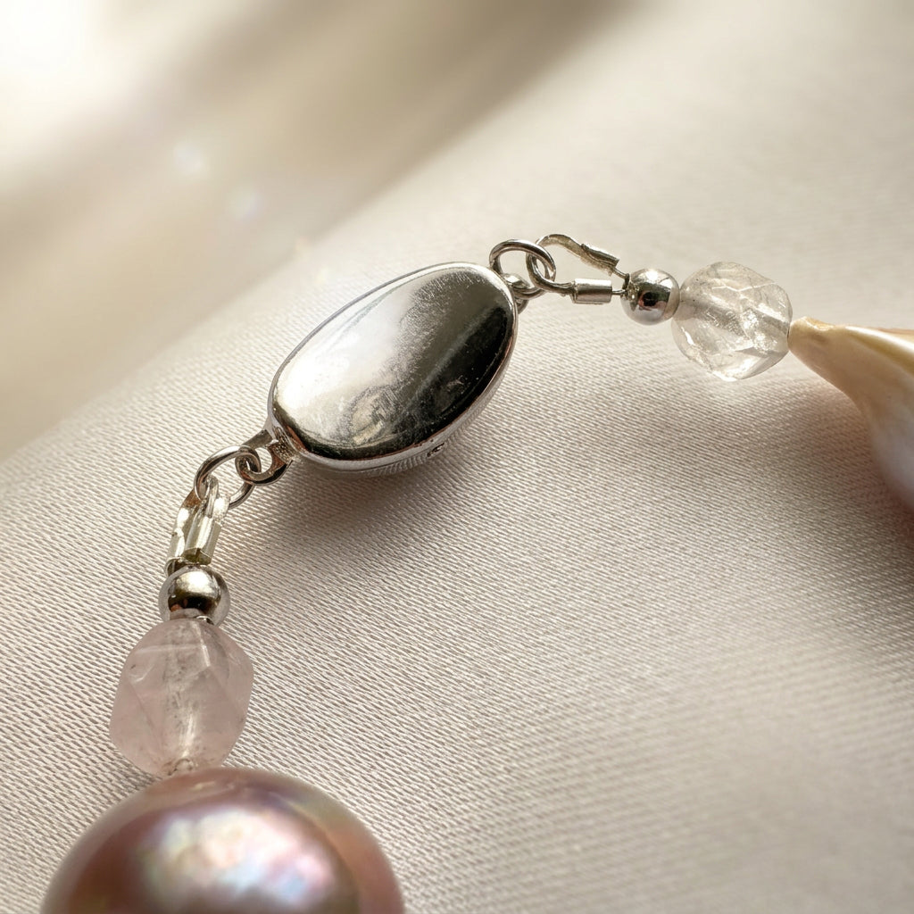 Close-up of a fastened oval sterling silver box clasp with a polished surface, beside faceted pale quartz beads, small silver accents and blush-toned keshi pearls on cream satin.