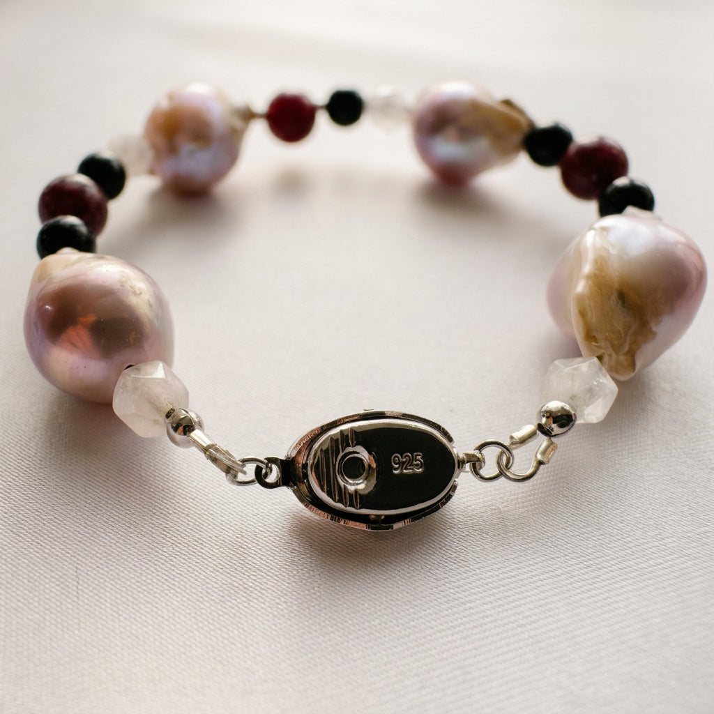 Keshi pearl bracelet arranged in an oval on cream satin, showing the underside of a fastened sterling silver box clasp stamped 925, with blush-cream pearls, faceted quartz, black onyx and deep red garnet beads.