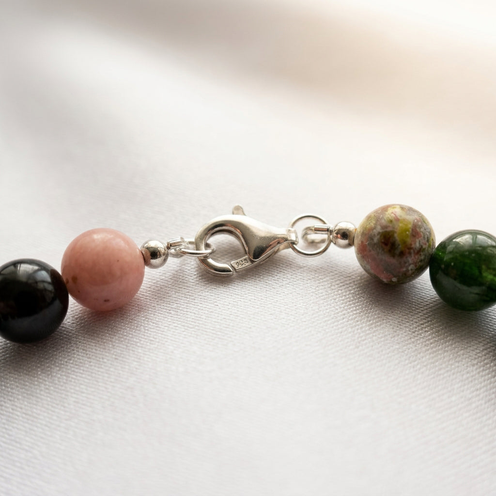 Close-up of a fastened sterling silver lobster clasp stamped 925, with connecting rings between pink, mottled earthy and deep green gemstone beads on cream fabric.