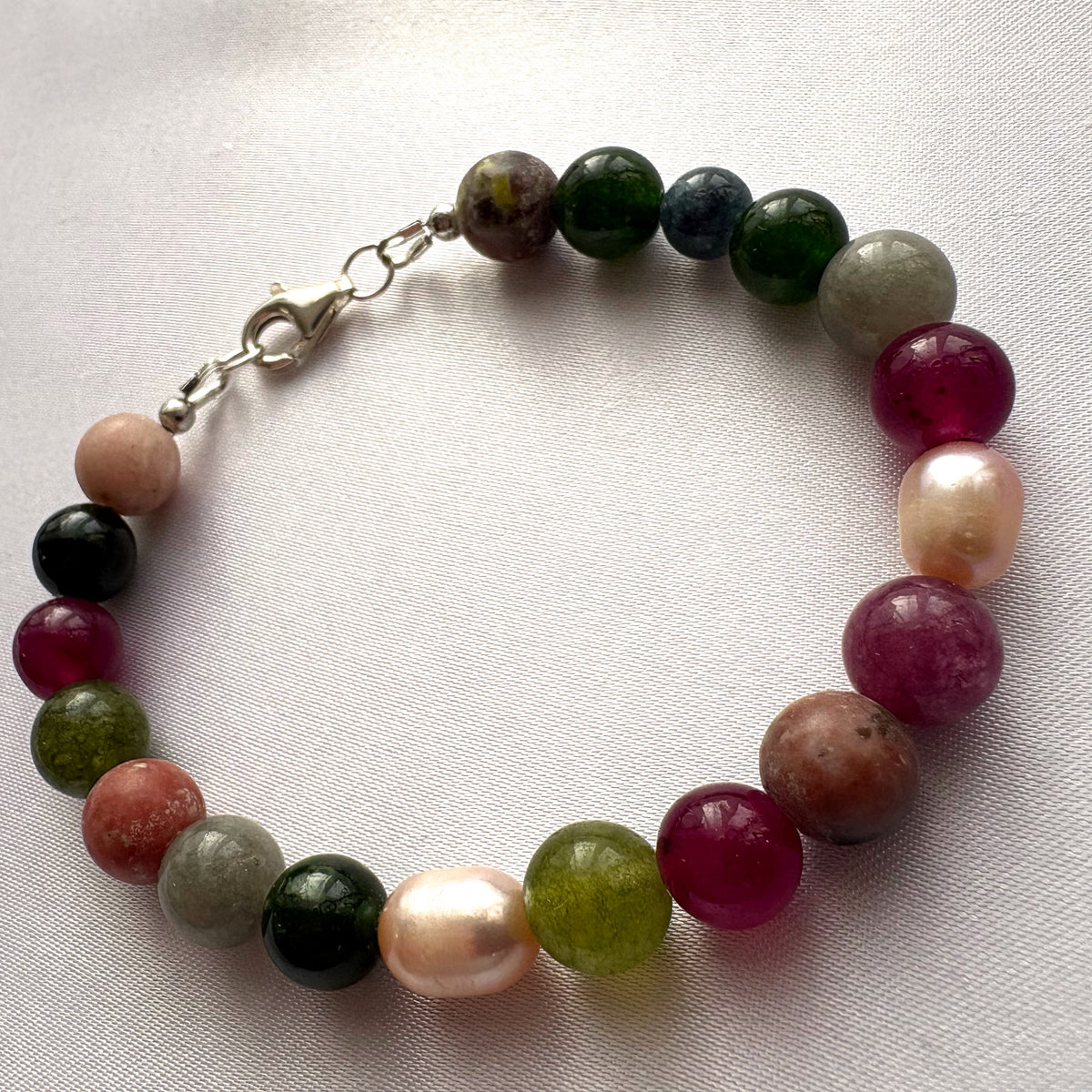 Jasper, tourmaline and baroque pearl bracelet arranged in a circle on white fabric, with magenta, olive-green, forest-green and earthy grey-brown beads, blush pearls and a sterling silver lobster clasp.