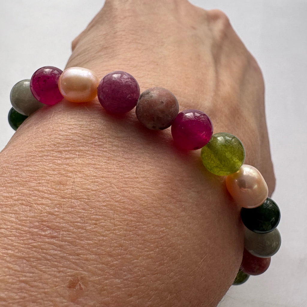 Angled close-up of a jasper, tourmaline and baroque pearl bracelet worn on a wrist, highlighting vivid magenta and translucent olive-green beads beside blush pearls and earthy grey-brown stones.