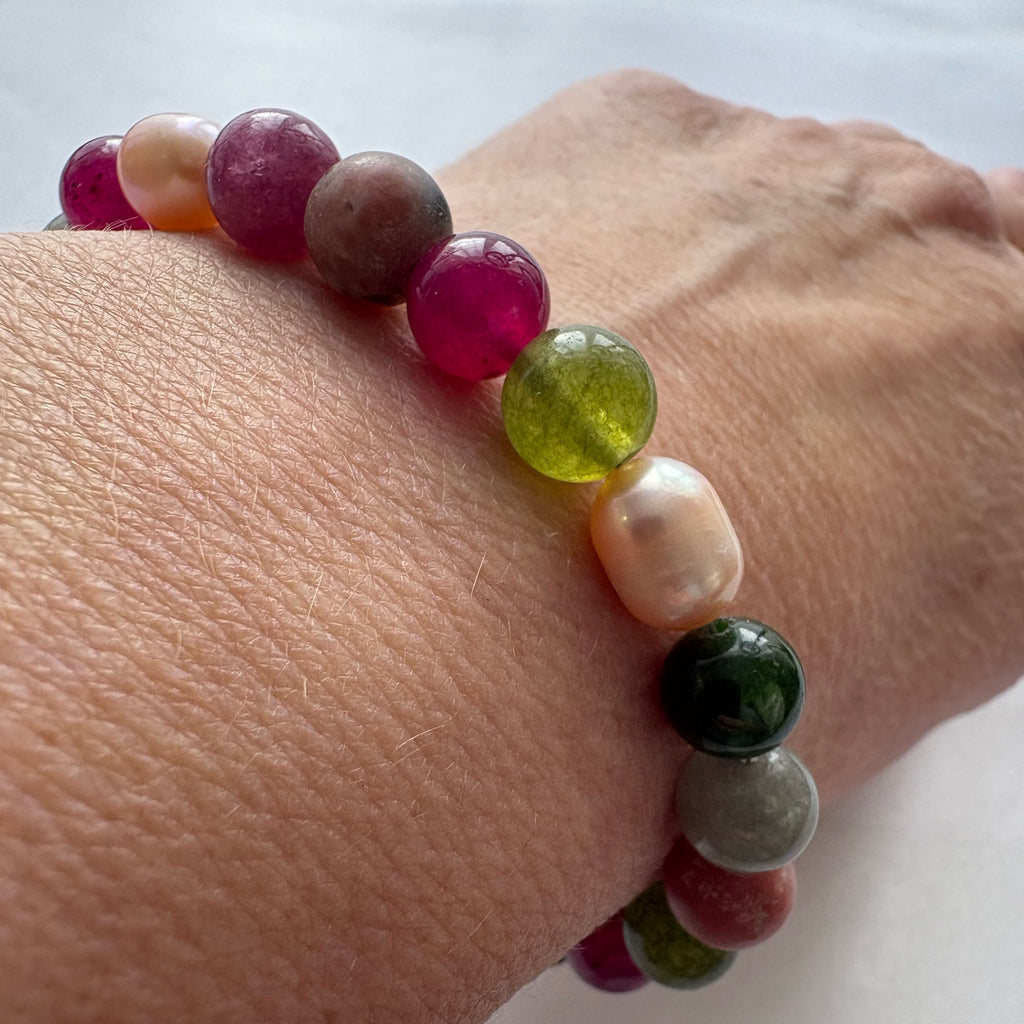 Close-up of a jasper, tourmaline and baroque pearl bracelet worn on a wrist, showing translucent magenta and olive-green beads beside a lustrous blush pearl, deep green stones and muted grey jasper.