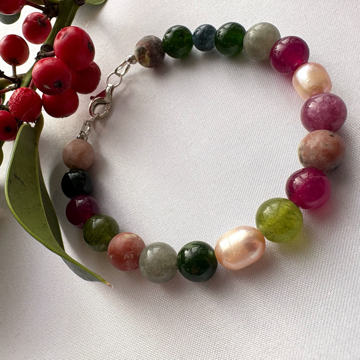 Jasper, tourmaline and blush baroque pearl bracelet arranged in a circle on white fabric beside red berries and green leaves, with a sterling silver lobster clasp.