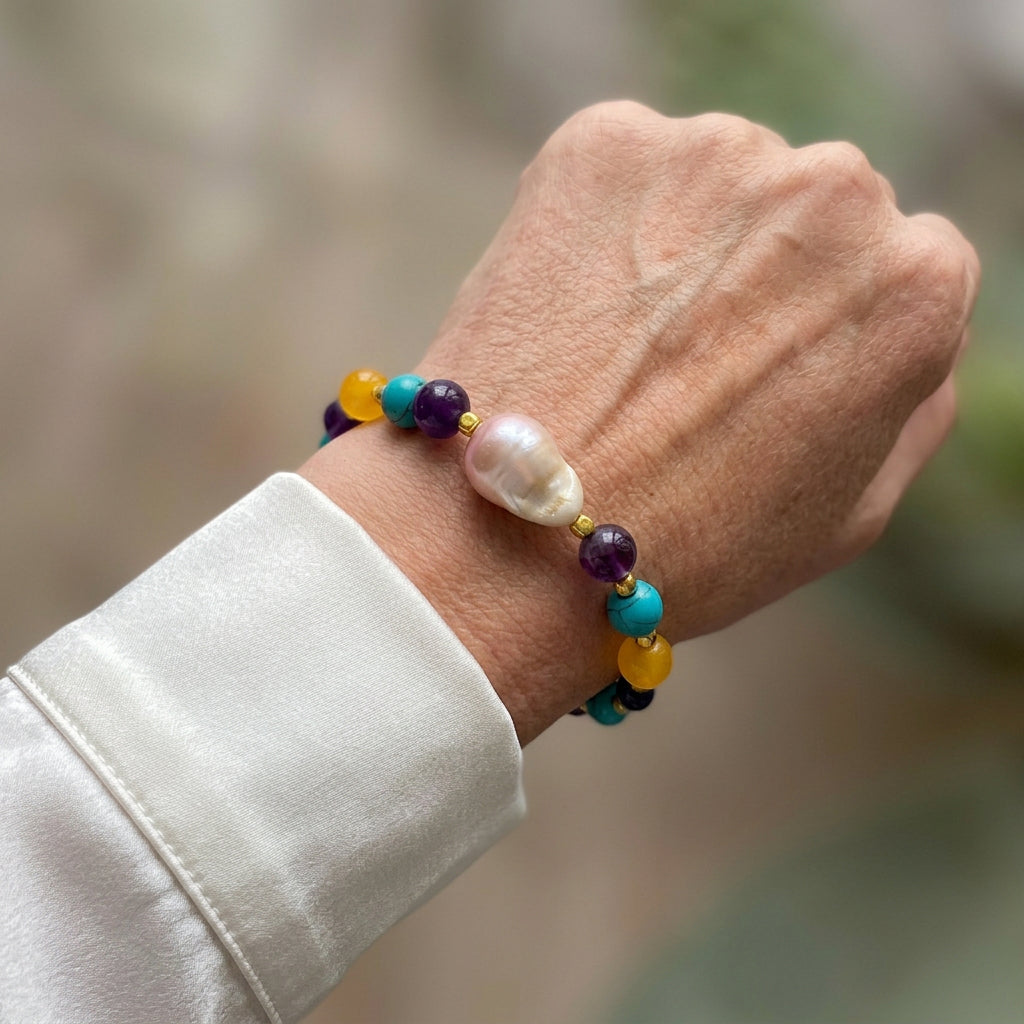 Bracelet with a large blush-cream keshi pearl, purple, turquoise-blue and yellow gemstone beads and gold-coloured spacers, worn on the wrist beside a white silk shirt cuff.