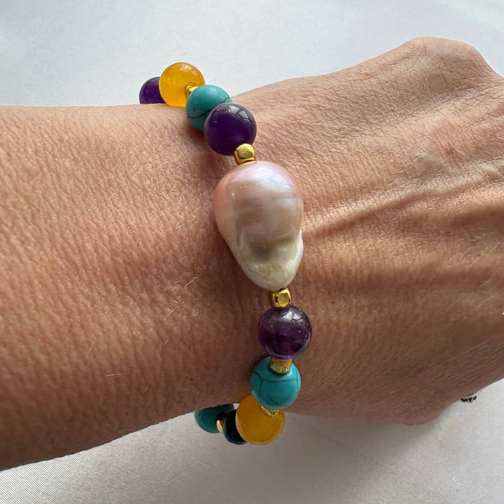 Close-up of a bracelet worn on a wrist, centred on a large blush-pink keshi pearl between purple amethyst beads, with teal-blue howlite, amber-yellow topaz and gold-plated spacers.