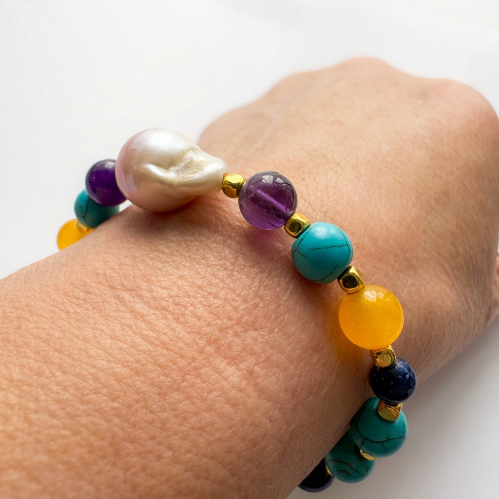 Angled close-up of a bracelet worn on a wrist, showing a blush-pink keshi pearl, translucent purple amethyst, veined teal-blue howlite and faceted amber-yellow topaz with gold-plated spacers.