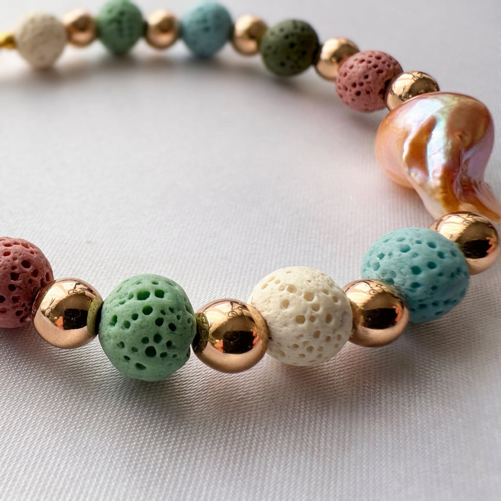 Close-up of porous mint-green, cream, teal and dusty pink lava beads alternating with polished gold-plated spacers, with an iridescent peach-gold keshi pearl on white fabric.⠋
