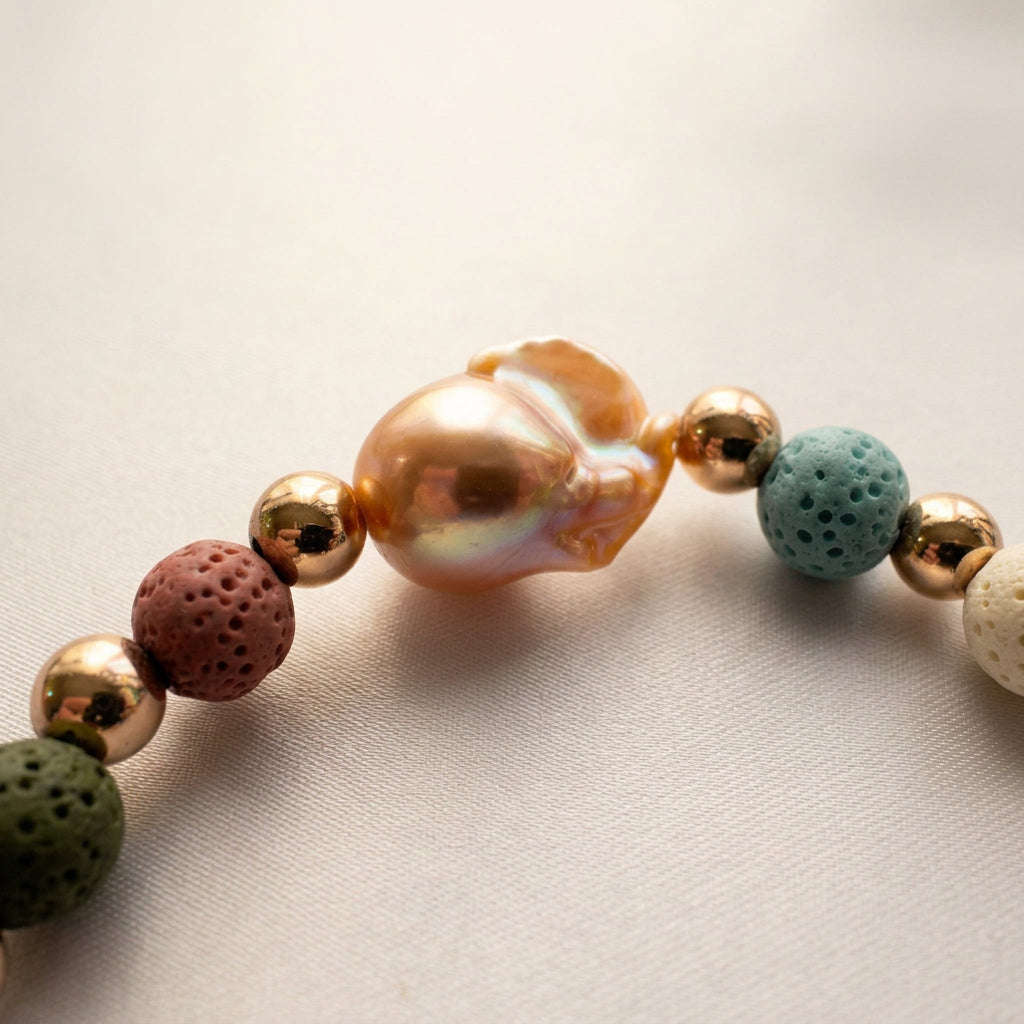 Close-up of a large peach-gold keshi pearl with rainbow iridescence between polished gold-plated spacers and porous dusty pink and teal lava beads on cream fabric.