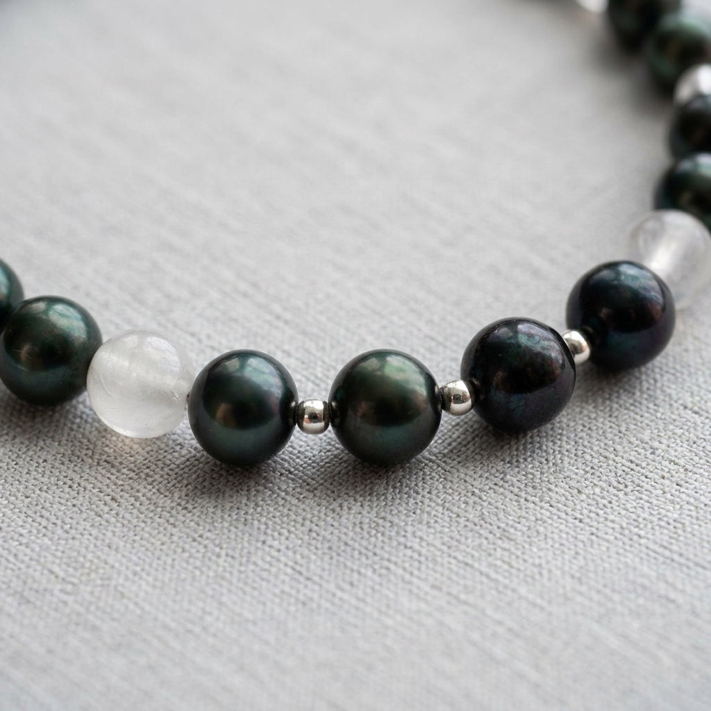 Close-up of peacock baroque pearls with green, teal and purple reflections, separated by small silver-coloured spacers and pale translucent gemstone beads on grey linen.